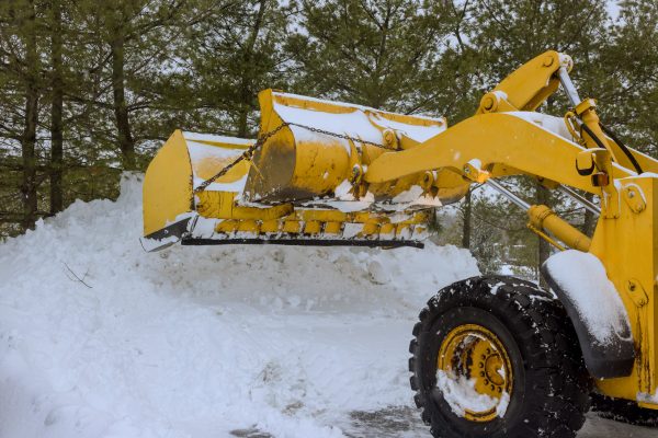 Heavy machinery works to remove thick snow buildup from driveway during winter storm in suburban area.